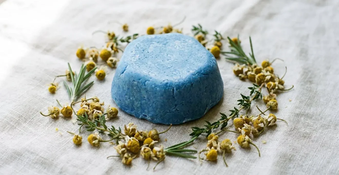 Barre de shampoing solide bleu posée sur une surface naturelle entourée de fleurs de camomille matricaire séchées
