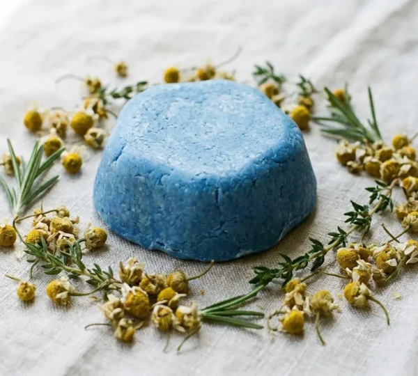 Barre de shampoing solide bleu posée sur une surface naturelle entourée de fleurs de camomille matricaire séchées